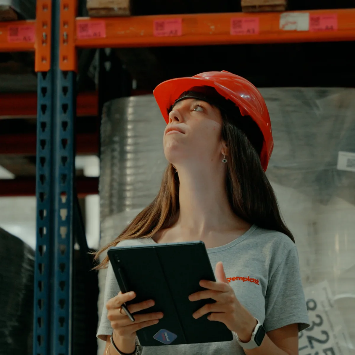Integrante del equipo de Gemplast, con casco de seguridad y remera de la marca, revisando el stock de los racks del depósito con una tablet
